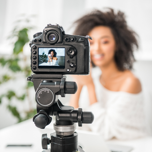 Fotocamera su treppiede riprende una giovane donna sorridente con capelli ricci, seduta davanti all’obiettivo mentre registra un video beauty.