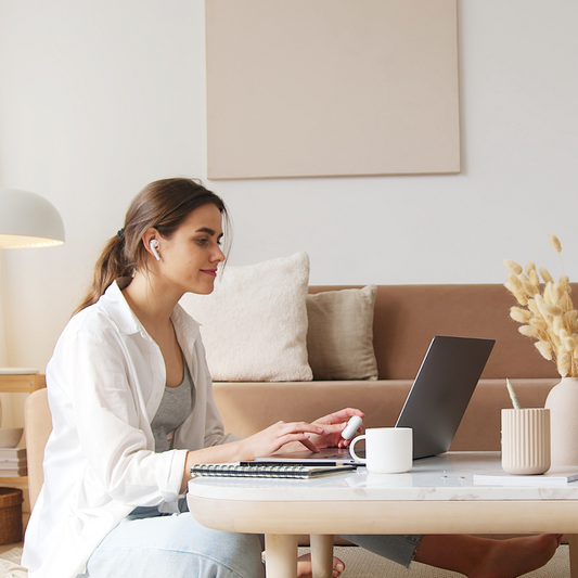 Donna sorridente seduta su un divano, lavora al laptop con auricolari, taccuino e tazza sul tavolino, in un ambiente domestico minimal e luminoso.