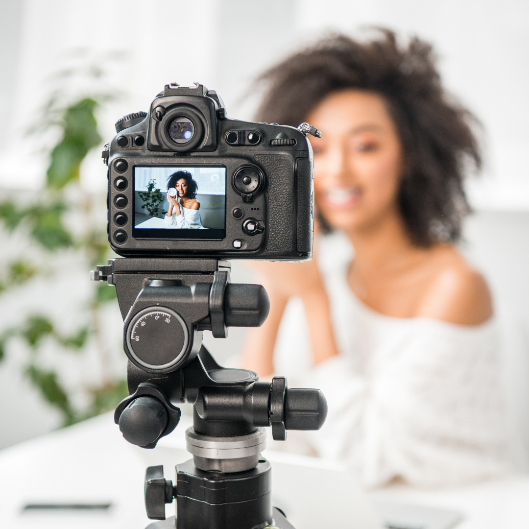 Fotocamera su treppiede riprende una giovane donna sorridente con capelli ricci, seduta davanti all’obiettivo mentre registra un video beauty.